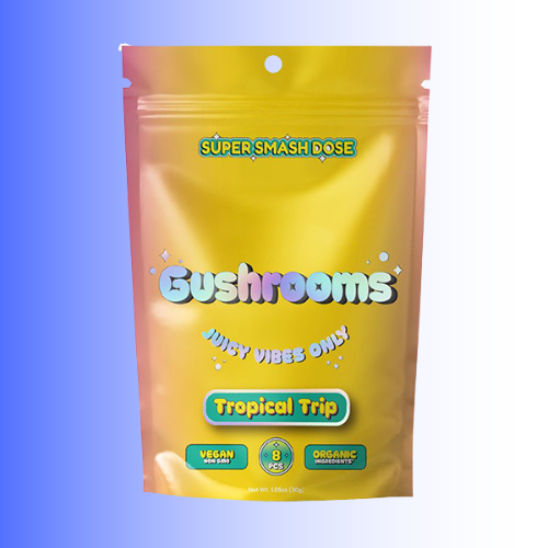 A yellow stand-up pouch of Gushrooms 'Tropical Trip Flavor' gummy candies, featuring colorful text and badges, set against a blue-to-white gradient background.
